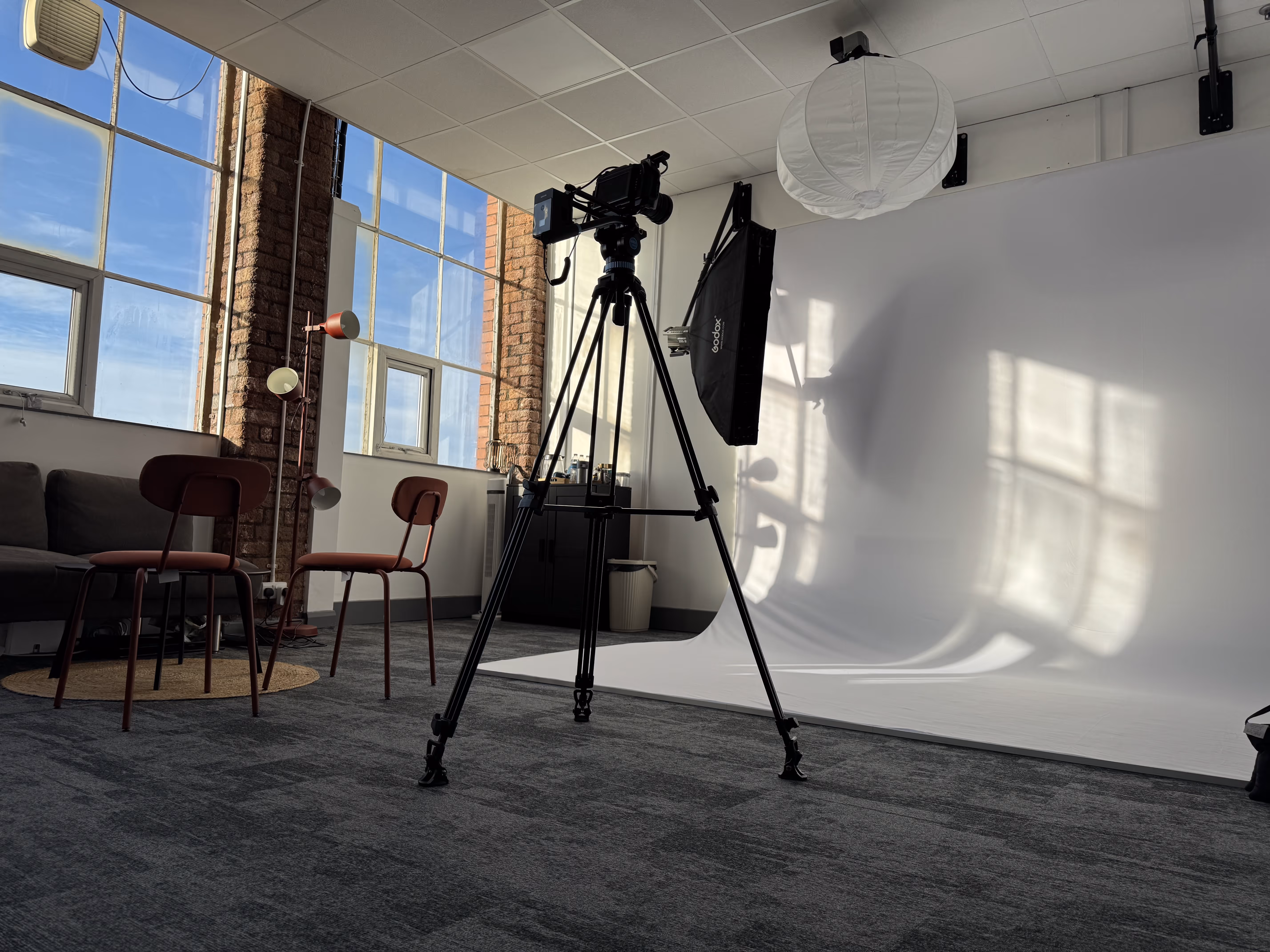 Photography studio with large windows, two chairs, a camera on tripod, softbox light, and white backdrop.
