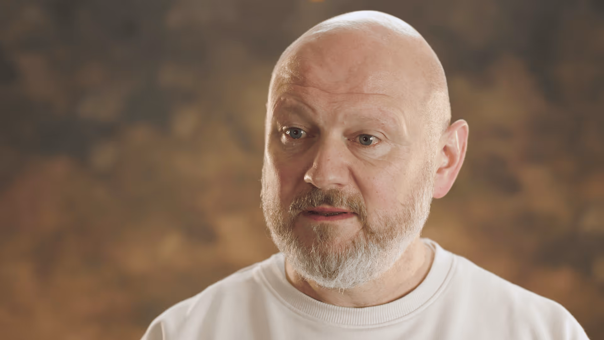 Close-up of a bald middle-aged man with a beard wearing a white shirt, speaking against a blurred brown background.