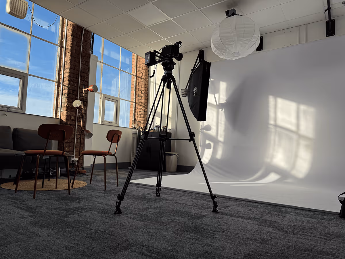 Photography studio with a camera on a tripod, two chairs, a couch, large windows, and a white backdrop with light and window shadows.